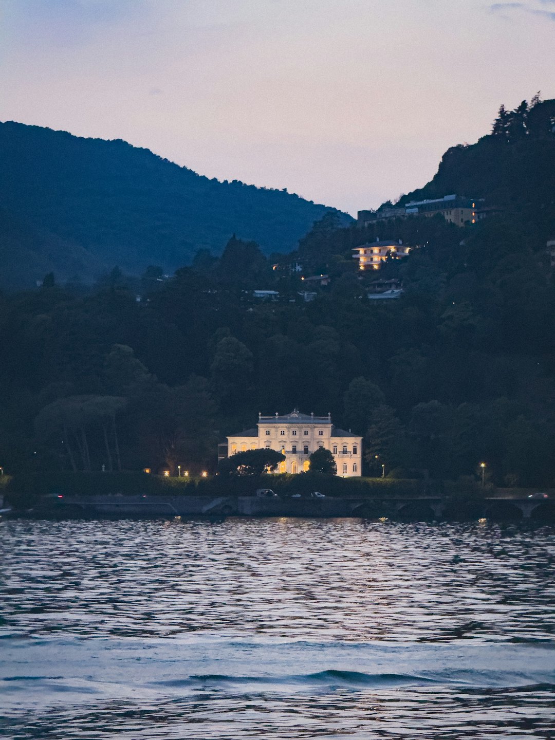 Eine beleuchtete Villa liegt in der Dämmerung am dunklen Ufer.